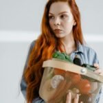 A young woman with red hair holding a basket of assorted fresh vegetables indoors.