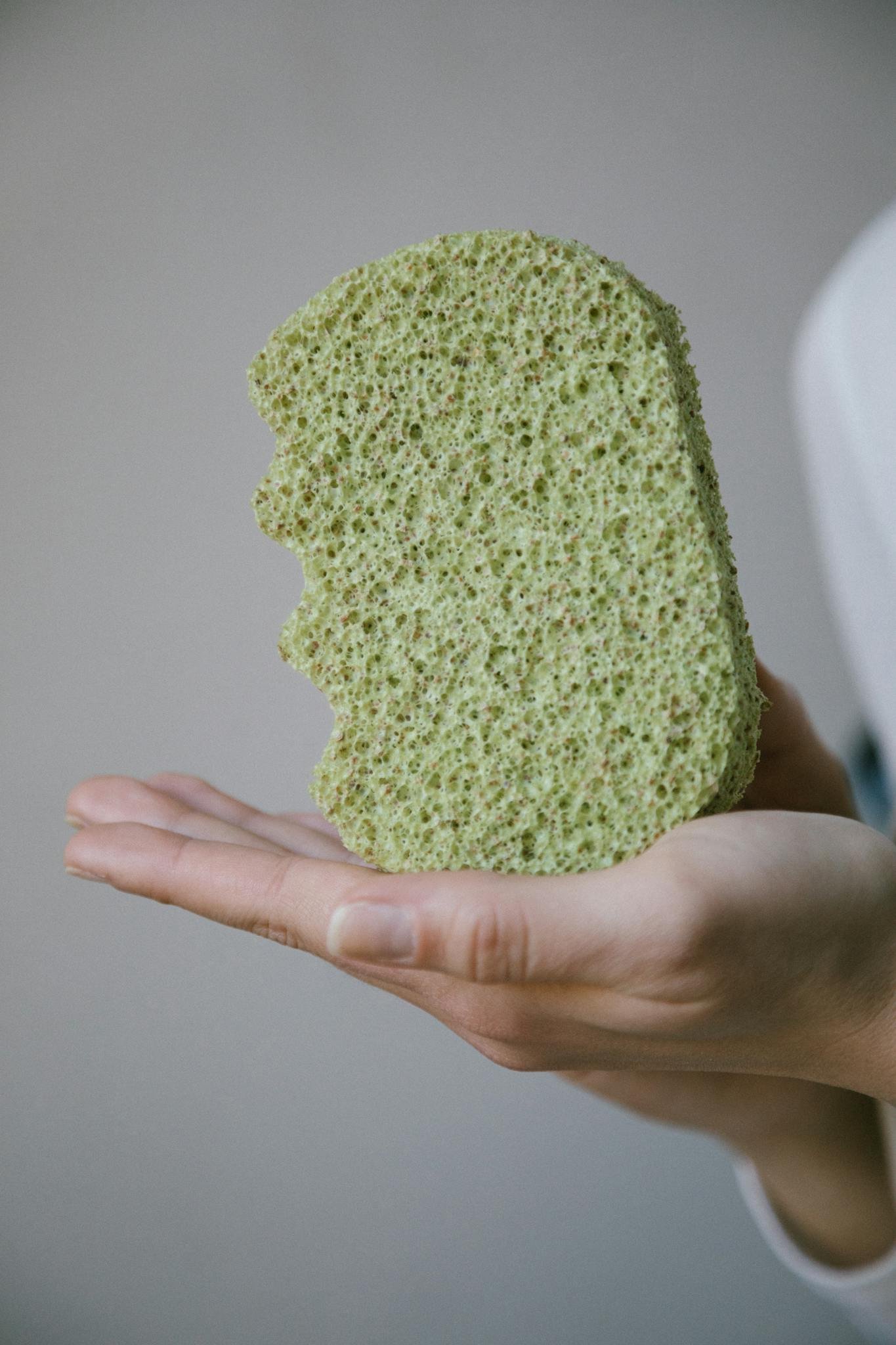 A green organic bath sponge held in a person's hand against a neutral background.